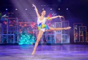 Eiskunstläuferin, die auf einem Bein steht und das andere Bein ausgestreckt hat, weißer Showanzug und bunter Gürtel